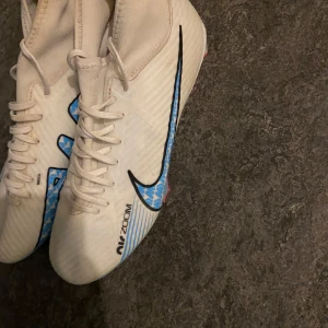 Nike Air Zoom fotbollsskor - Snygga vita Nike Air Zoom fotbollsskor med blå och rosa detaljer. Skorna har snörning och en hög ankelkrage för extra stöd. Perfekta för fotbollsplanen med en modern design och bra grepp.