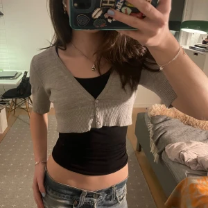 Grå glittrig bolero - Säljer en snygg grå glittrig bolero med trekvartsärm och knappar framtill. Koftan är croppad och har en ribbad kant som ger en fin passform.