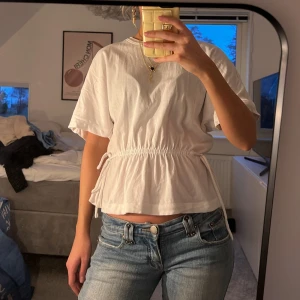 Vit kortärmad topp med knytband - Säljer en vit kortärmad topp med knytband i midjan. Toppen har en avslappnad passform och är perfekt för en casual look. Den är tillverkad i ett mjukt material som känns skönt mot huden.