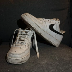 Nike Air Force i vitt och svart - Säljer ett par klassiska Nike Air Force sneakers i vitt med svart swoosh. Skorna har snörning och är tillverkade i skinn. Perfekta för en stilren look.