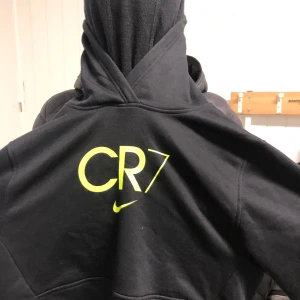 Svart hoodie från Nike med CR7-logga - Säljer en svart hoodie från Nike med CR7-logga i gult på ryggen. Den har en stor Nike-swoosh i silver på framsidan. Perfekt för alla CR7-fans!