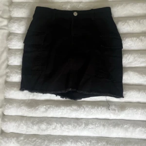 Svart jeans kjol från H&M - Snygg svart jeanskjol från H&M med praktiska fickor på sidorna. Kjolen har en knapp och dragkedja framtill för enkel påklädning. Perfekt för en stilren look.