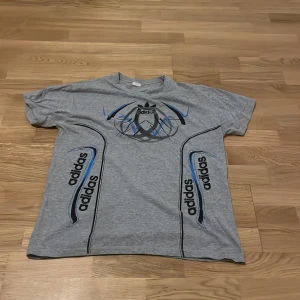 Grå t-shirt från Adidas - Säljer en grå t-shirt från Adidas med tryck i svart och blått. T-shirten har korta ärmar och ett sportigt mönster med Adidas-loggan framtill. Perfekt för en avslappnad stil.