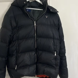 Svart dunjacka från GANT - Säljer en svart dunjacka från GANT med huva och dragkedja. Jackan har två framfickor med dragkedjor och en snygg logga på bröstet. Insidan är fodrad i en kontrasterande orange färg. Perfekt för kyliga dagar! Priset kan diskuteras!