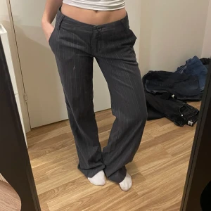 Grå lågmidjade randiga kostymbyxor - Snygga grå kostymbyxor med diskreta ränder. Byxorna har en klassisk design med knappar och dragkedja framtill. Passar en som är runt 170-175cm i längden. Jag är 160cm för referens 