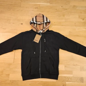 Burberry hoodie  - Säljer en stilren svart hoodie från Burberry med klassiskt rutigt mönster i huvan. Tröjan har dragkedja och två fickor framtill. Perfekt för en avslappnad och trendig look.
