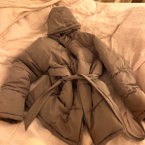 Beige pufferjacka med bälte - Säljer en snygg beige pufferjacka med bälte och dragkedja. Jackan har en huva och långa ärmar, perfekt för kyliga dagar. Den är både stilren och praktisk, vilket gör den till ett måste i garderoben. Passar perfekt för höst och vinter.