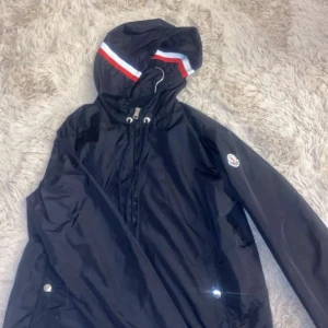 Svart vindjacka från Moncler - Snygg svart vindjacka från Moncler med huva och dragkedja. Jackan har en stilren design med Moncler-logga på ärmen och röd-vit randdetalj på huvan. Perfekt för blåsiga dagar. NFC Scan finns 