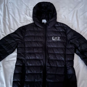 Svart pufferjacka från Emporio Armani - Snygg svart pufferjacka från Emporio Armani med EA7-logga på bröstet. Köpt från butik och säljer på grund av att den inte används. Passar perfekt för vår och höst. Om det är något du undrar över är der bara att skicka ett meddelande 