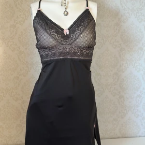 Svart spetsklänning med rosa detaljer - Elegant svart klänning med spetsdetaljer och rosa rosetter. Klänningen har smala axelband och en v-ringad spetsöverdel som ger en feminin touch. Postar inte med instabox 