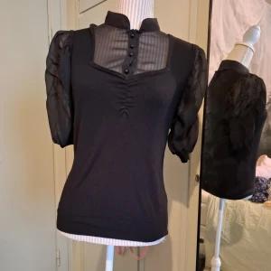 Svart blus från Next - Elegant svart blus från Next med transparenta detaljer och knappar framtill. Blusen har korta puffärmar och en åtsittande passform. Perfekt för en stilren look. Saknar en knapp men går att ersättas