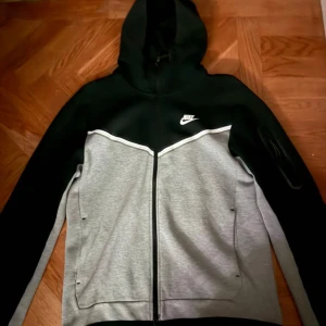 Svart och grå hoodie från Nike - Säljer en snygg svart och grå hoodie från Nike med dragkedja och huva. Tröjan har en sportig look med två fickor framtill och en liten Nike-logga på bröstet. Perfekt för en casual stil. Använd fåtal gånger