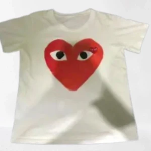 Vit t-shirt med rött hjärta från CDG - Säljer en vit t-shirt med ett stort rött hjärta med ögon på framsidan. Perfekt för en avslappnad stil med en rolig twist. Passar bra till jeans eller shorts.