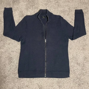 Mörkblå kofta från Carl William - Säljer en stilren mörkblå kofta från Carl William. Den har en praktisk dragkedja framtill och är tillverkad i ett mjukt stickat material. Perfekt för kyligare dagar med sin ribbade krage och muddar.