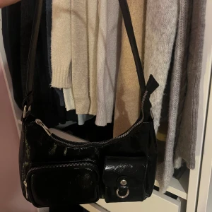 Svart axelväska med dragkedja - Snygg svart axelväska i syntetmaterial med praktiska dragkedjor och en stilren design. Väskan har en justerbar axelrem och flera fack för enkel organisering. 