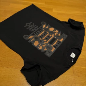 Svart t-shirt från Burberry  - Snygg svart t-shirt från Burberry med ett stort tryck i grått och orange på framsidan. Perfekt för en stilren look med en touch av lyx. T-shirten har en klassisk passform och korta ärmar.