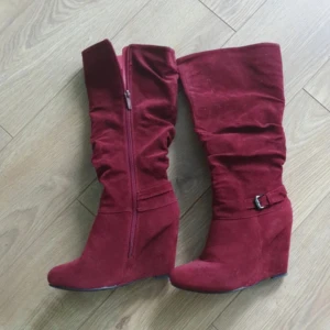 Vinröda stövlar med kilklack - Snygga vinröda stövlar i mocka med kilklack och dragkedja på sidan. De har en dekorativ rem med spänne 💘