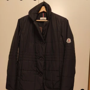 Svart dunjacka från Moncler - Säljer en stilren svart dunjacka från Moncler med knappar framtill. Jackan har en quiltad design och långärmad passform. Perfekt för kyligare dagar med sin varma och bekväma stil.