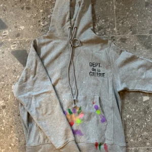 Grå hoodie med färgstänk - Säljer en grå hoodie från Dept. de la Galerie med färgstänk i olika färger på fickan. Tröjan har en klassisk design med huva och dragsko samt en stor magficka. Perfekt för en avslappnad stil.