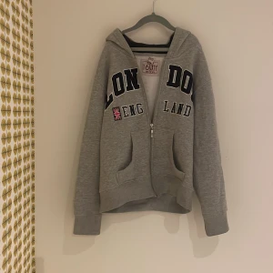 Grå hoodie med London-tryck - Säljer en grå hoodie från Toasty med texten 'London England' och en liten brittisk flagga på bröstet. Tröjan har dragkedja och en bekväm huva. Perfekt för en avslappnad stil.