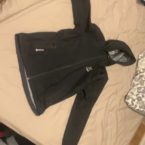 Svart vindjacka från Haglöfs Gore tex - Säljer en svart vindjacka från Haglöfs med dragkedja och huva. Jackan är tillverkad i GORE-TEX-material och har två praktiska fickor med dragkedjor. Perfekt för utomhusaktiviteter och skyddar mot vind och regn.