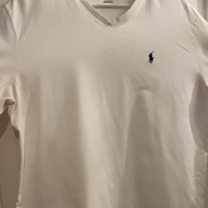 Vit t-shirt från Ralph Lauren - Säljer en klassisk vit t-shirt från Ralph Lauren med v-ringning och den ikoniska loggan broderad i blått på bröstet. Perfekt för en stilren och avslappnad look.