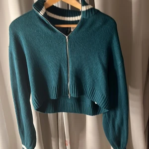 Grön ribbad hoodie med dragkedja - Säljer en snygg grön ribbad hoodie med dragkedja framtill. Den har en hög krage med vita ränder och långa ärmar. Perfekt för en avslappnad stil. Passar bra till jeans eller leggings.