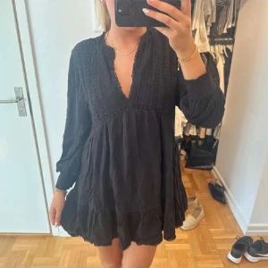 Klänning - Snygg svart klänning med v-ringning och spetsdetaljer. Den har långa ärmar och en luftig passform som ger en avslappnad look. Perfekt för både vardag och festliga tillfällen.