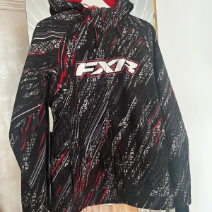 Svart och röd jacka från FXR - Säljer en cool svart jacka från FXR med röda och vita detaljer. Jackan har ett mönster med FXR-loggor och en dragkedja framtill. Perfekt för kyliga dagar med sin långa ärmar och huva.
