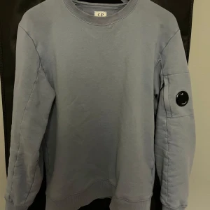 Ljusblå  sweatshirt från C.P. Company - Säljer en stilren ljusblå sweatshirt från C.P. Company. Tröjan är i bra skick. Tyvärr är det några riktigt små vita fläckar vid kragen men inget som syns tydligt. Sällan använd därav säljer jag den.