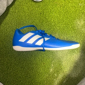 Blå fotbollsskor från Adidas - Säljer ett par snygga blå fotbollsskor från Adidas i mycket bra skick. De har vita detaljer och snörning för perfekt passform. Perfekta för inomhusfotboll eller träning. Skorna är bekväma och ger bra grepp på planen.
