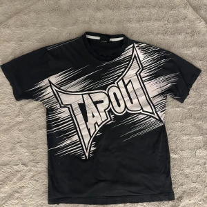 Svart T-shirt från Tapout - Säljer en svart T-shirt från Tapout i mycket bra skick. Den har ett stort, vitt Tapout-tryck på framsidan och ett mindre på baksidan. Perfekt för träning eller vardagsbruk. Materialet känns mjukt och bekvämt. Passar dig som gillar en sportig stil!