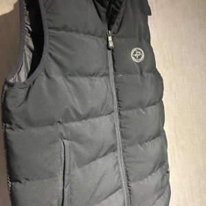 Svart dunväst från Pelle P - Säljer en snygg svart dunväst från Pelle P i storlek M. Västen har en dragkedja framtill och två praktiska fickor. Perfekt för höst och vår när du vill hålla dig varm utan att bära en jacka. Den är i mycket bra skick och redo för nya äventyr!