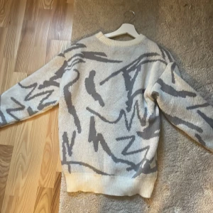 Beige och grå stickad tröja - Säljer en supermysig stickad tröja i beige och grått med ett abstrakt mönster. Perfekt för kyliga dagar! Tröjan har långa ärmar och ribbade muddar vid ärmslut och nederkant. Den är i bra skick och passar perfekt för höst och vinter.