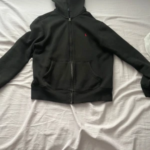 Svart hoodie från Ralph Lauren - Säljer en snygg svart hoodie från Ralph Lauren i storlek L i barn passar på någon som är 160 cm.Den har en dragkedja framtill och en klassisk röd logga på bröstet. Perfekt för höst och vinter, och den är supermjuk och bekväm. Passar både till vardags och chillkvällar. 🖤