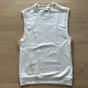 Vit ärmlös tröja från Weekday - Säljer en stilren vit ärmlös tröja från Uniqlo. Perfekt för lager-på-lager eller som den är under varmare dagar. Tröjan har en V-ringning och är i ett mjukt bomullsmaterial. Passar både till vardags och mer uppklädda tillfällen.