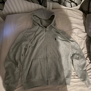 Grå hoodie med dragkedja - Säljer en snygg och bekväm grå hoodie från Polo Ralph Lauren. Den har en dragkedja framtill och en klassisk logga på bröstet. Perfekt för både vardag och chill. Storleken är inte angiven, men den ser ut att vara normal passform. Hör av dig vid intresse! Pris diskuteras storleken är L men sitter som M