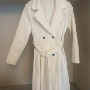 Vit kappa med bälte - Säljer en elegant vit kappa i mycket bra skick. Den har en klassisk design med dubbelknäppning och ett matchande bälte i midjan. Perfekt för höst och vinter! Långärmad och i ett stilrent snitt som passar både till vardags och fest.
