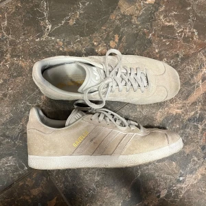 Beige Adidas Gazelle sneakers - Säljer ett par snygga beige Adidas Gazelle sneakers i mocka. De är i bra skick men med tecken på användning, speciellt framme vid tårna. 