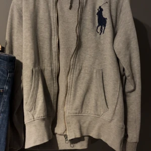 Grå hoodie från polo ralph lauren - Säljer en snygg grå hoodie från Ralph Lauren med det klassiska logotypen broderad i blått på bröstet. Den har en dragkedja framtill och en skön huva. Perfekt för en avslappnad stil eller för att hålla sig varm under kyliga dagar. Passar till både jeans och joggers!