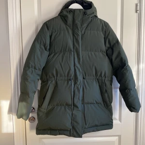 Mossgrön dunjacka från Peak Performance - Säljer en grön dunjacka från Peak Performance i storlek S. Jackan är perfekt för kalla vinterdagar med sin långa design och huva. Den har en dragkedja framtill och är i mycket bra skick. Perfekt för både vardag och utomhusaktiviteter!