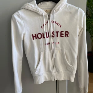 Vit hoodie från Hollister - Säljer en vit hoodie från Hollister med röd text på bröstet. Den har en dragkedja och är i bomullsmaterial. Perfekt för en avslappnad stil, men har en liten fläck på framsidan. Passar bra till både höst och vår.💖