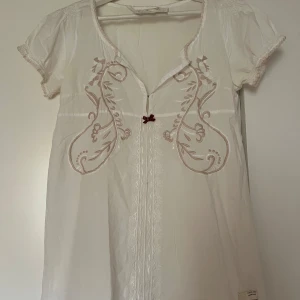 Vit blus från Odd Molly - Säljer en superfin vit blus från Odd Molly med vackra broderade detaljer i beige. Blusen har korta ärmar och en söt liten rosett framtill. Perfekt för vår och sommar! Passar både till vardags och festliga tillfällen.
