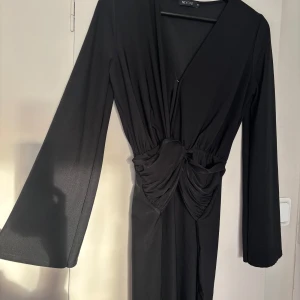 Svart långklänning från Nelly  - Elegant svart långklänning från Nelly med v-ringning och långa ärmar. Klänningen har en snygg drapering i midjan som ger en smickrande passform. Perfekt för festliga tillfällen eller en kväll ute. Materialet känns mjukt och bekvämt mot huden.