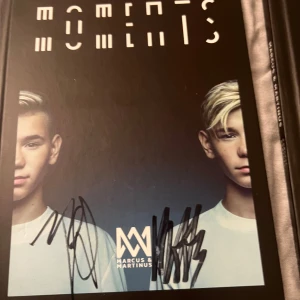 Moments - Säljer boken 'Moments' av Marcus & Martinus. Omslaget är svart med deras porträtt och titeln i vit text. Boken är signerad av artisterna, vilket gör den extra speciell för fans. Perfekt för samlare eller som present till någon som älskar deras musik!