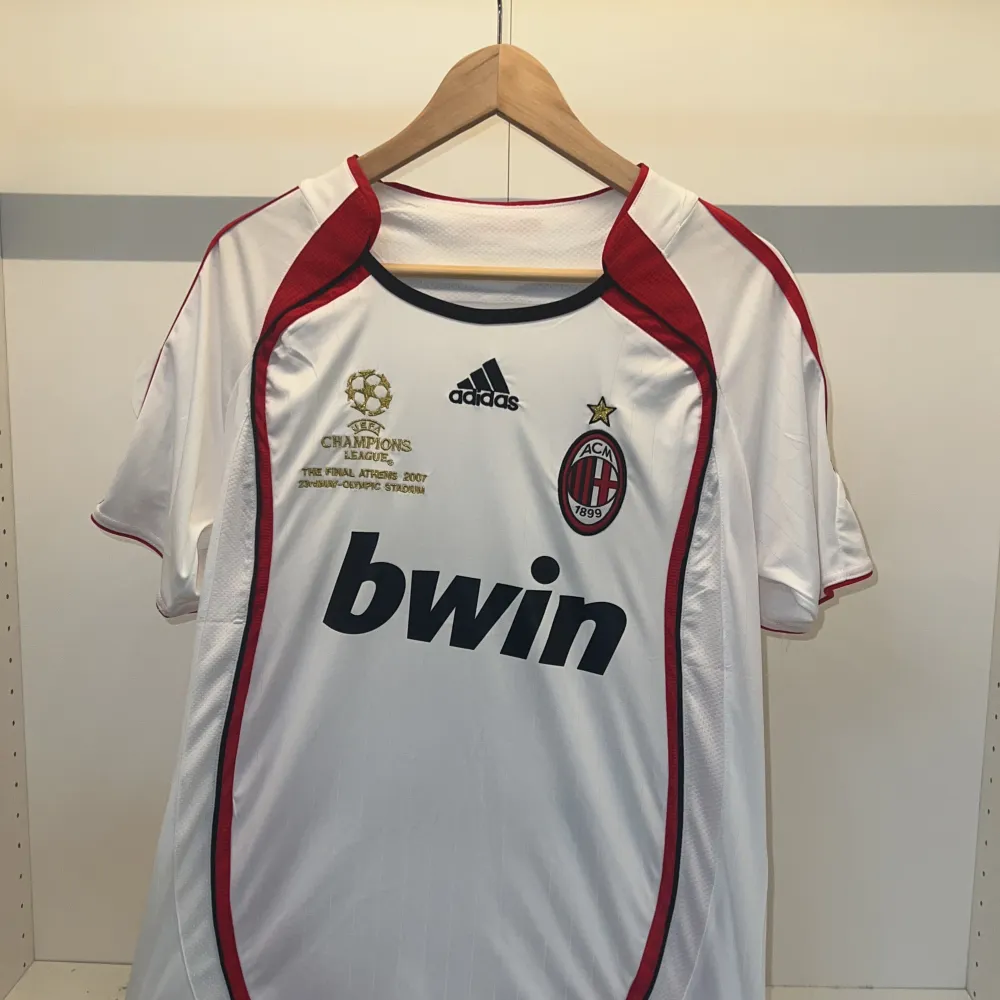 Säljer en vit AC Milan fotbollströja från Adidas med Kaka' och nummer 22 på ryggen. Tröjan har korta ärmar och är dekorerad med röda och svarta detaljer. Den har Champions League-märken och Bwin som sponsorlogga. Perfekt för samlare eller fans av Kaka'! ⚽. T-shirts.