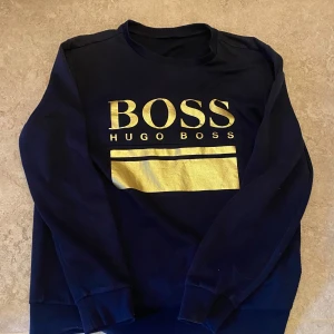 Svart tröja från Hugo Boss - Säljer en snygg svart tröja från Hugo Boss med guldtryck på bröstet. Tröjan har långa ärmar och är perfekt för både vardag och fest. Den är i bra skick och passar perfekt till höst och vinter. Skön och stilren design som funkar till det mesta!