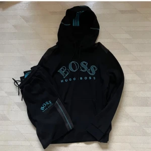 Svart hoodie och byxor från Hugo Boss - Säljer en snygg svart hoodie från Hugo Boss med stort BOSS-tryck på framsidan. Den har en klassisk passform med justerbar huva och ribbade muddar. Perfekt för en avslappnad stil eller träning. Passar både höst och vinter.