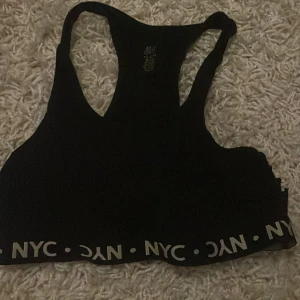 Svart sporttopp från H&M - Säljer en svart sporttopp från H&M i storlek M. Den har en elastisk kant med texten 'NYC' runtom. Perfekt för träning eller en aktiv dag. Toppen är i bra skick och redo för nya äventyr!