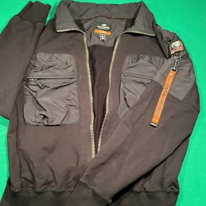 Svart bomberjacka från Parajumpers - Säljer en snygg svart fleece/ cardigan från Parajumpers i storlek M men passar S. Kort för mig (175cm) men bra annars. Jackan har en cool patch på ärmen och en orange dragkedjedetalj. Den är i fleece och har praktiska fickor med dragkedja. Perfekt för höst och vår! Väldigt fint skick. Nypris runt 3500kr. Hör av dig vid frågor och funderingar, mvh Christian 